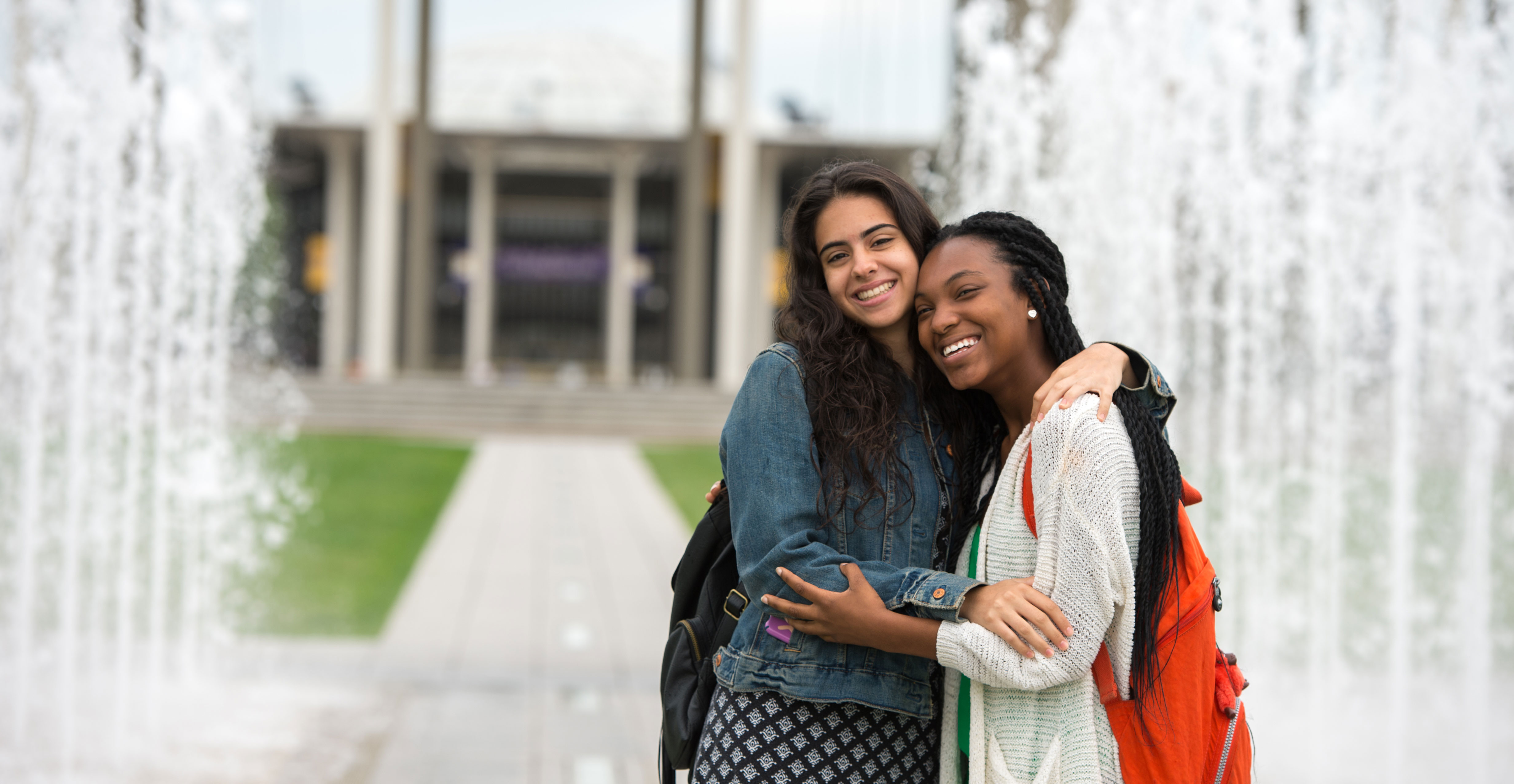 Two students wearing casual clothing and backpacks pose for a photo, embracing and smiling while standing in front of a fountain.