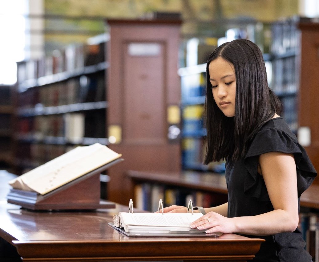 Female student in the UAlbany Downtown Library reading from a large ringed binder.