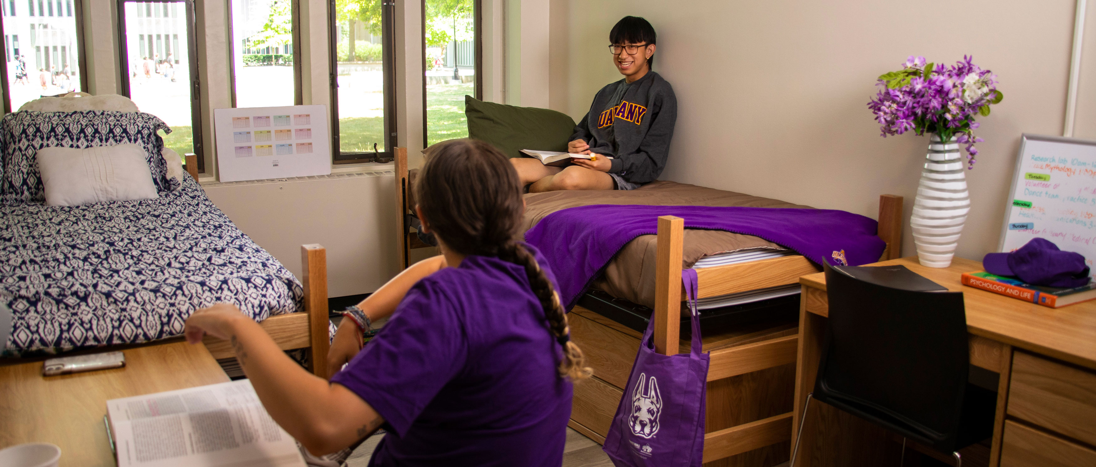 A student sitting on a twin bed smiles at their roommate, who is faced away from the camera and sitting at a desk next to another bed inside a residence hall.