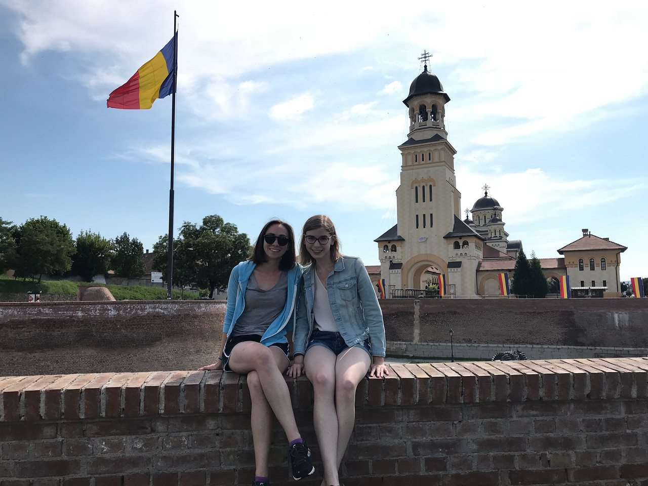 Two students sit on a wall with a Romanian building and flag in the background.