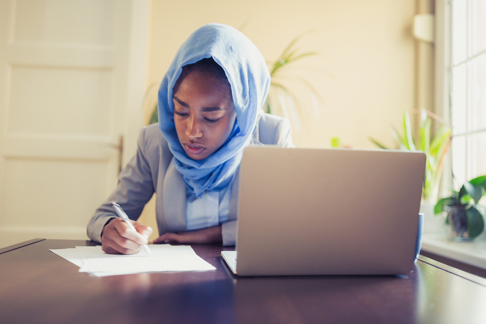A woman sits at a table with a laptop in front of her. She uses paper to take notes.