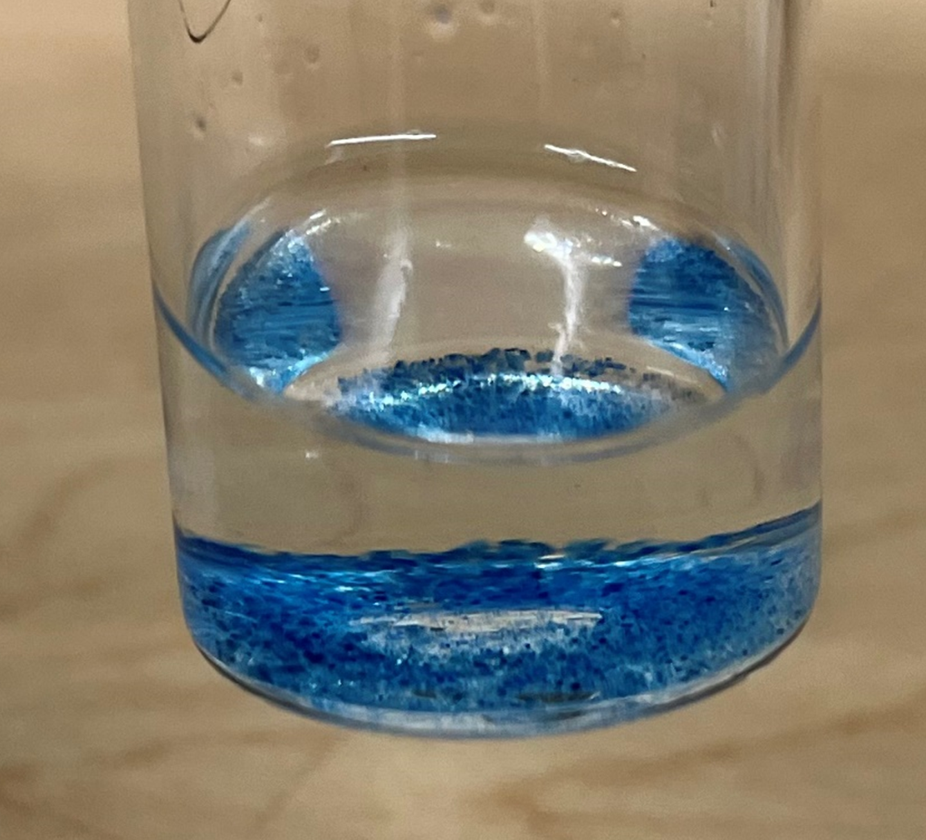 A glass vial containing clear liquid with bright blue particles settled at the bottom.