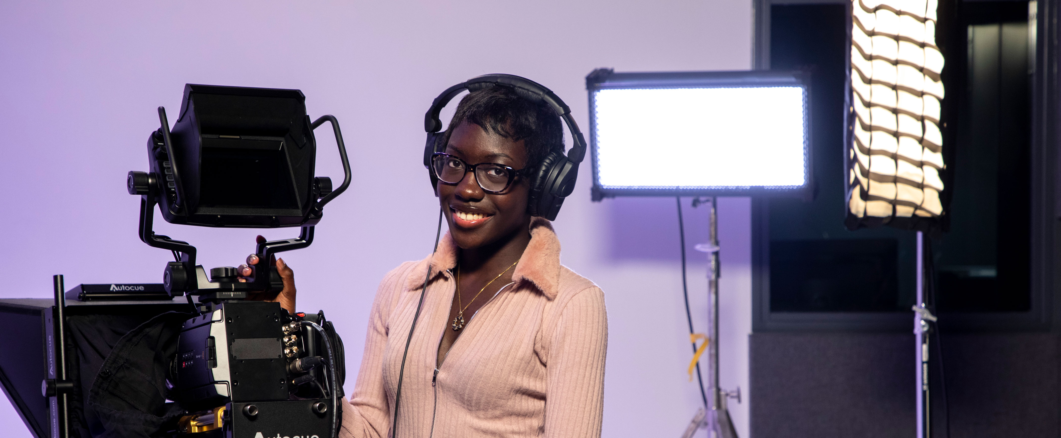 A student wearing a pink blouse and headphones smiles and poses for a photo with camera equipment, including a teleprompter, camera and lighting gear.
