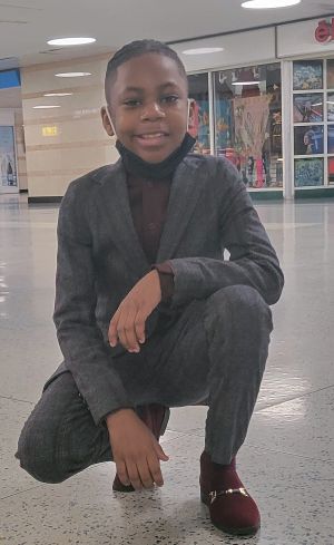 A young boy, wearing a gray suit and cranberry-colored shoes, squats in concourse