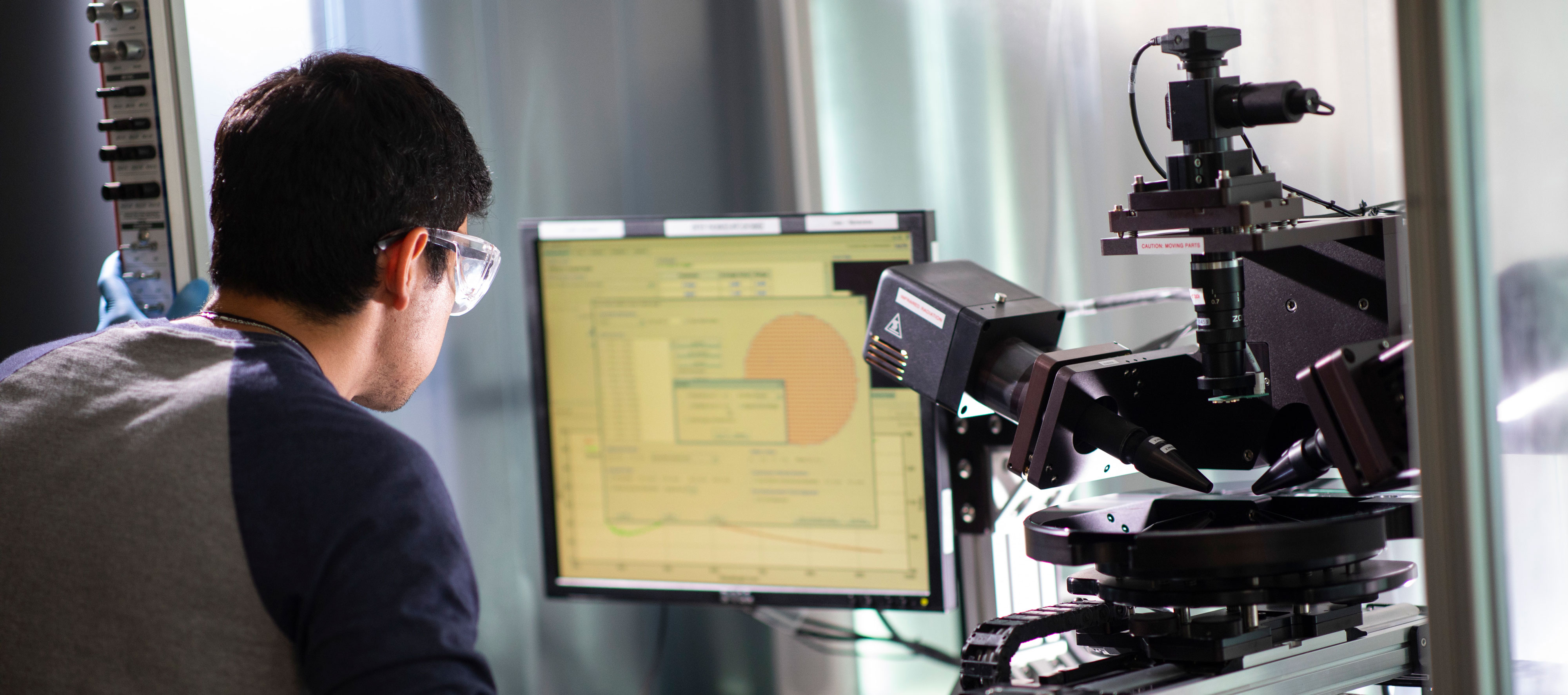 A researcher in safety glasses leans over a computer situated beside research equipment.