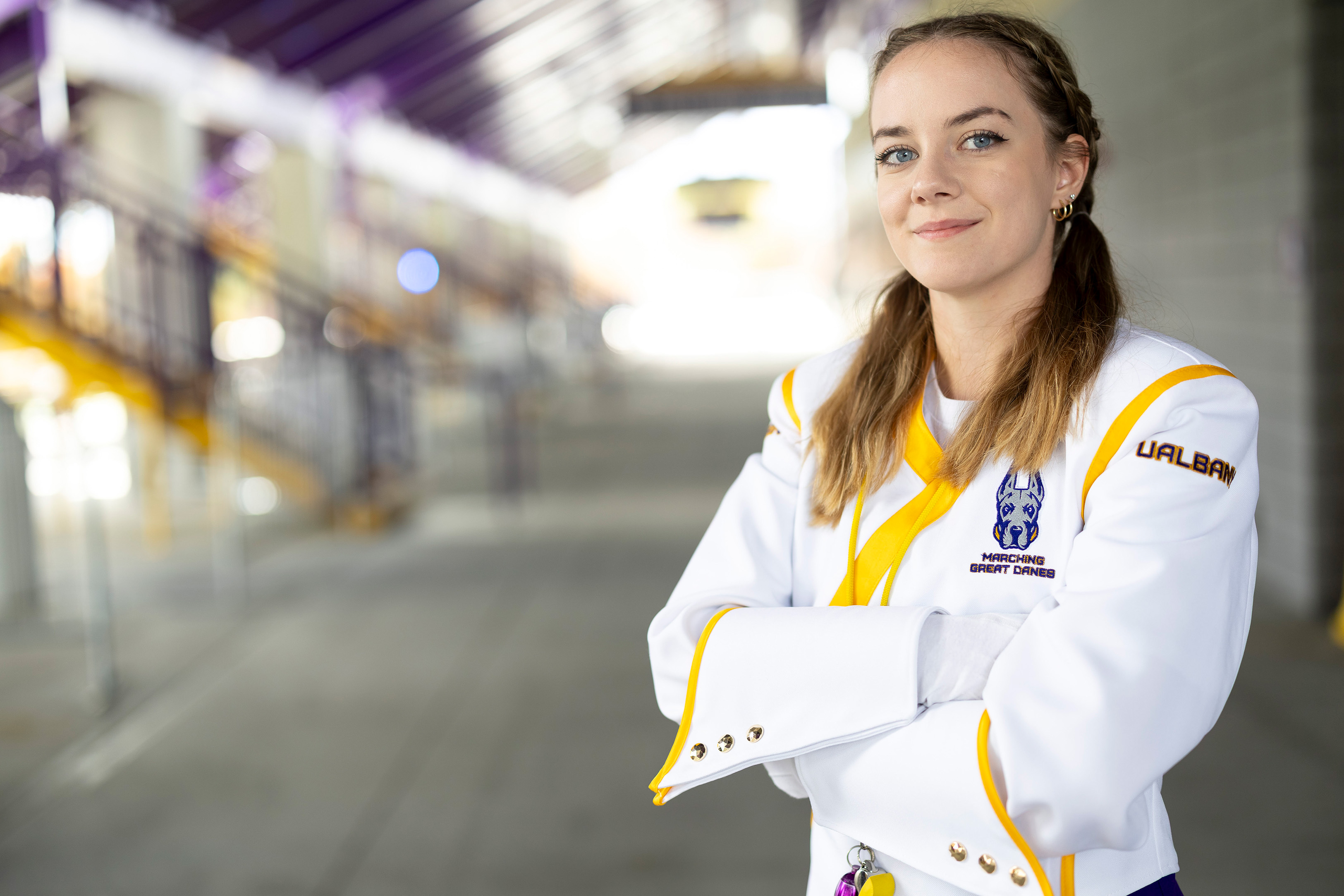 A UAlbany Drum Major poses with their arms crossed, smiling at the camera professionally. They are wearing the white coat of the UAlbany Marching Band Uniform with gold and purple accents.