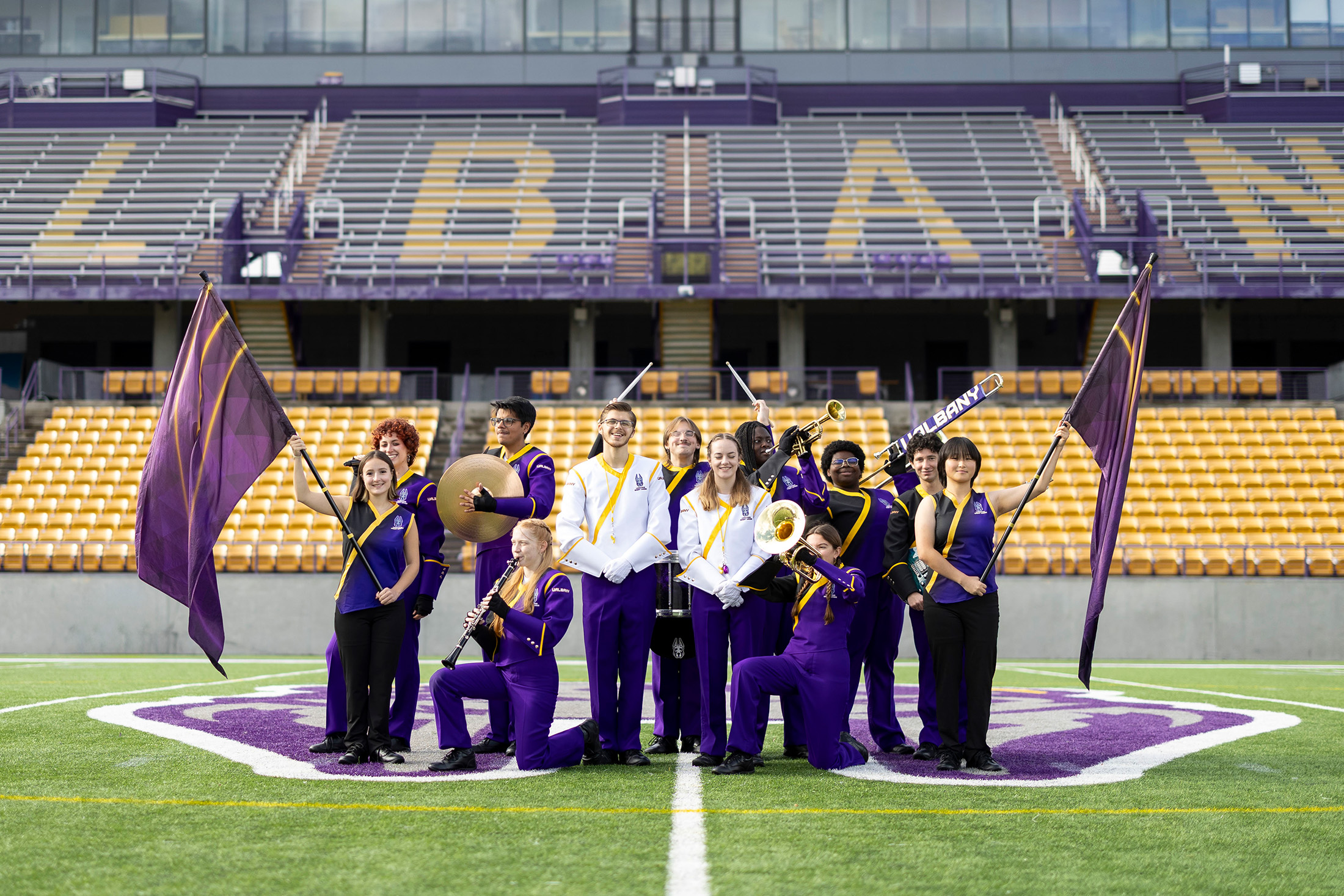 An ensemble of marching band members, two colorguard with flags, two drum majors, two percussionists, a clarinetist, and two trumpet players pose for a group photo in the UAlbany Football stadium. They are all wearing the UAlbany marching band uniform, which is mostly purple with gold accents. The drum majors' uniforms have purple pants with a white coat and gold accents.