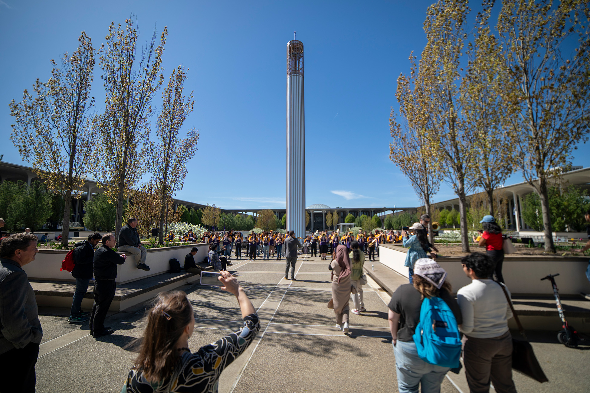 The UAlbany marching band performs in front of the carilon tower with a group of onlookers enjoying the performance under a beautiful blue sky.