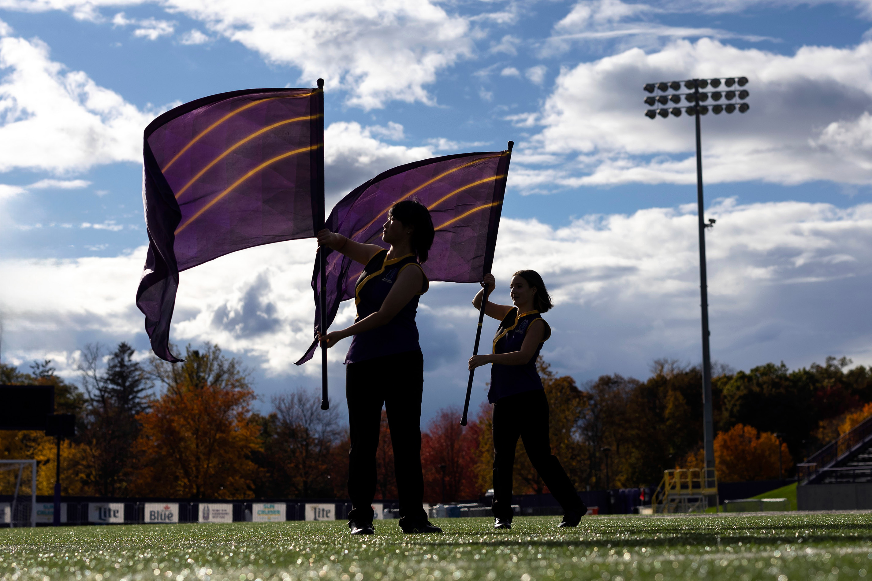 Two members of the UAlbany Color Guard raise purple and gold flags under a clear blue sky while standing on a field of green grass.