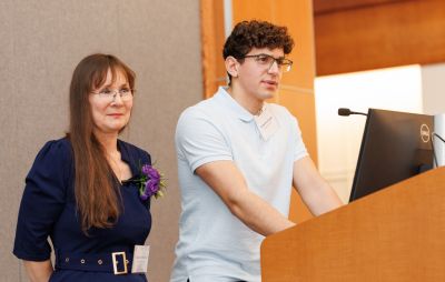 A smiling woman stands beside a young man speaking at a lectern.