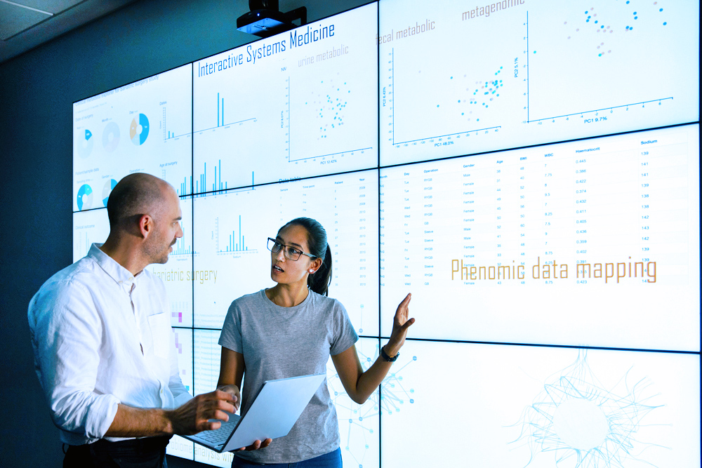 Two professionals viewing multiple screens of data charts and graphs.