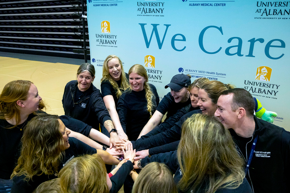 A group of people forming a hand stack in front of a We Care sign in the UAlbany gym.