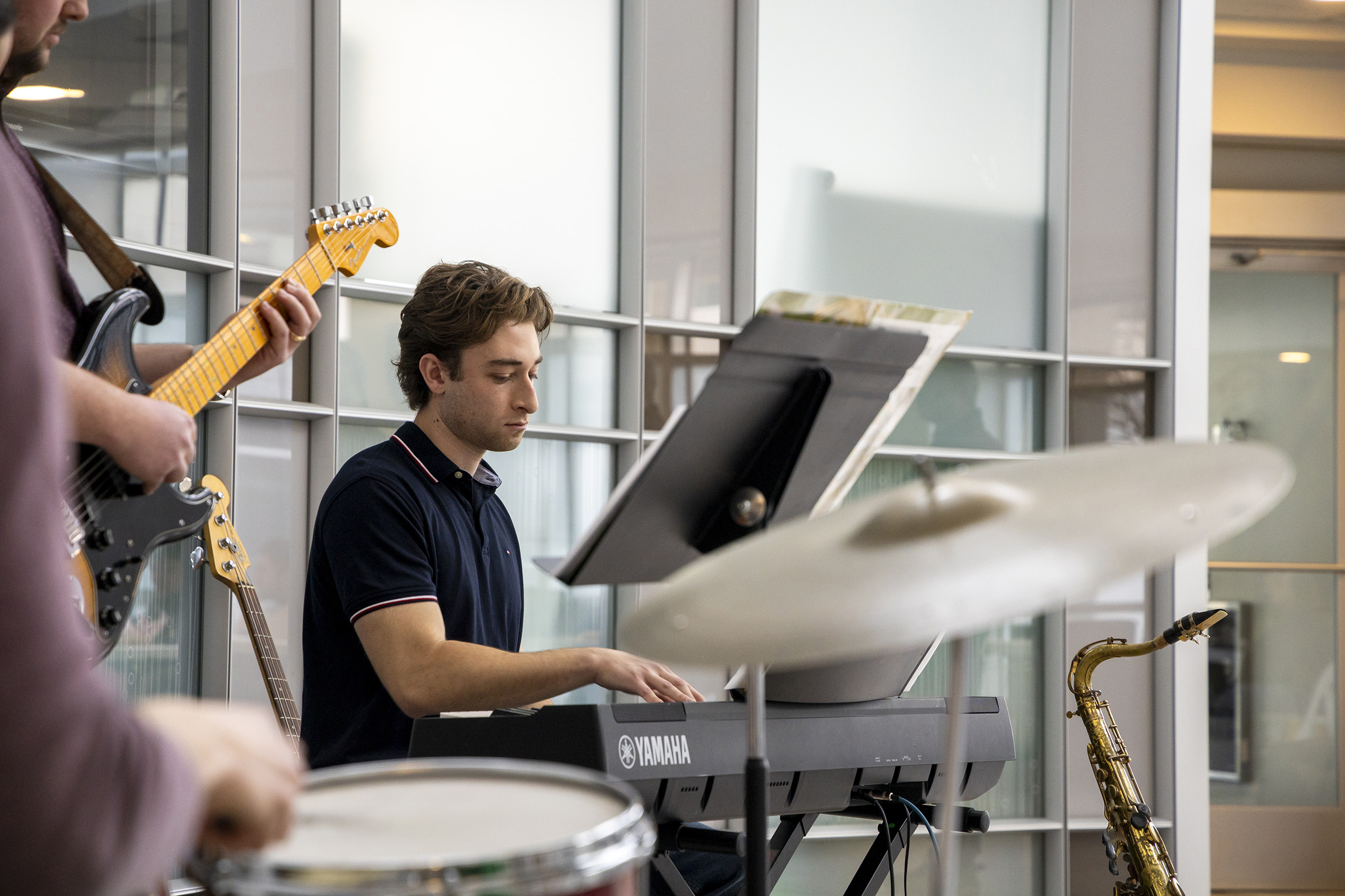 A young man wearing a black shirt plays a keyboard while accompanied by a guitar and drums inside of a glass rotunda.