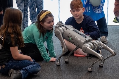 Young children gather around and interact with a robotic dog