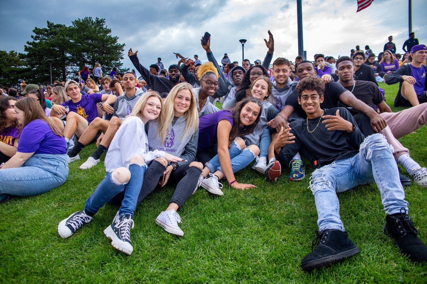 A dozen students smile and pose for a photo while sitting on a grassy hill above UAlbany's football stadium during a home game.