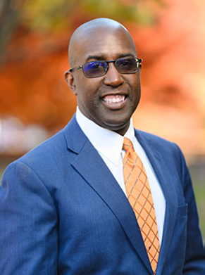 A man in sunglasses, an orange tie and blue suit smiles for a portrait outdoors