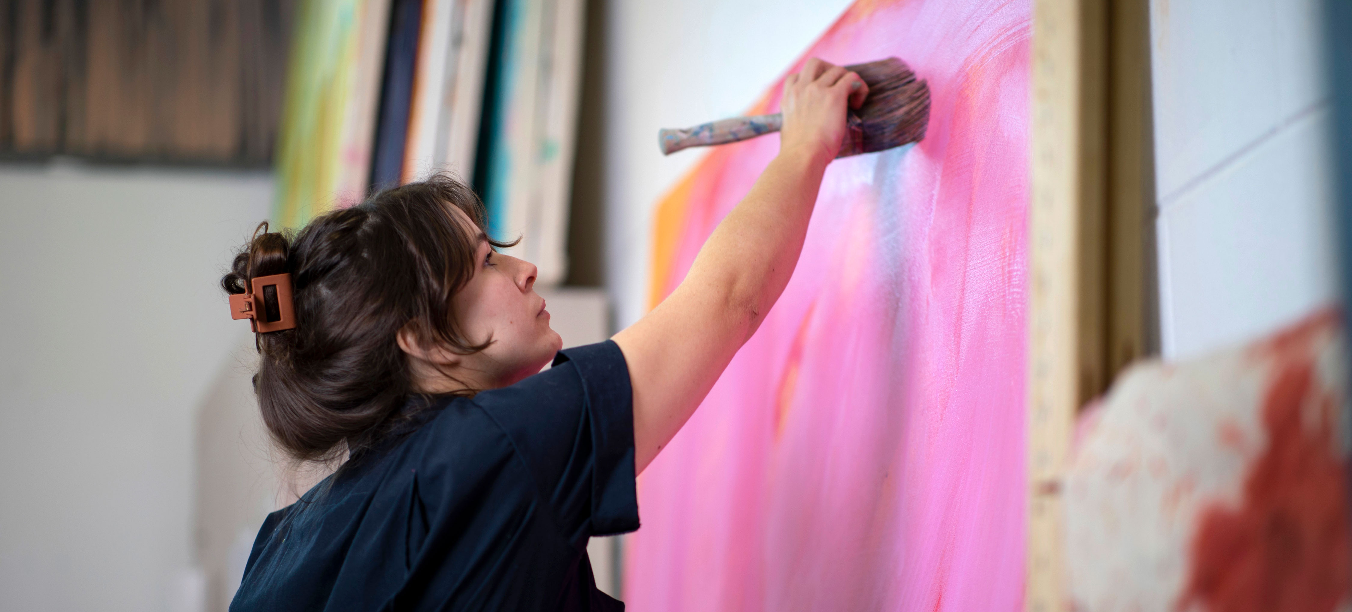 A student wearing navy coveralls uses a large paintbrush to add pink paint to a large canvas inside an art studio.