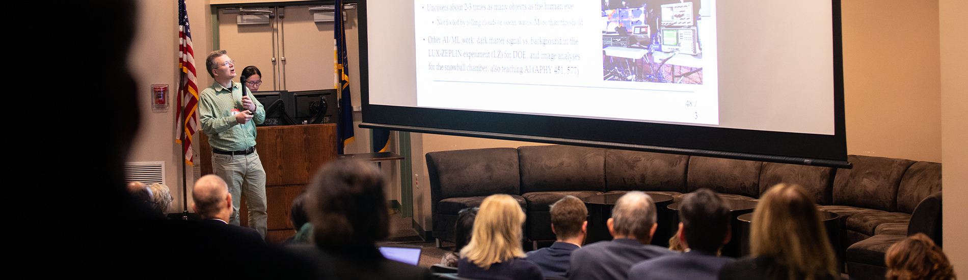 A professor in a green button up and khakis holds a microphone and stands in front of a presentation screen while speaking to a crowded auditorium