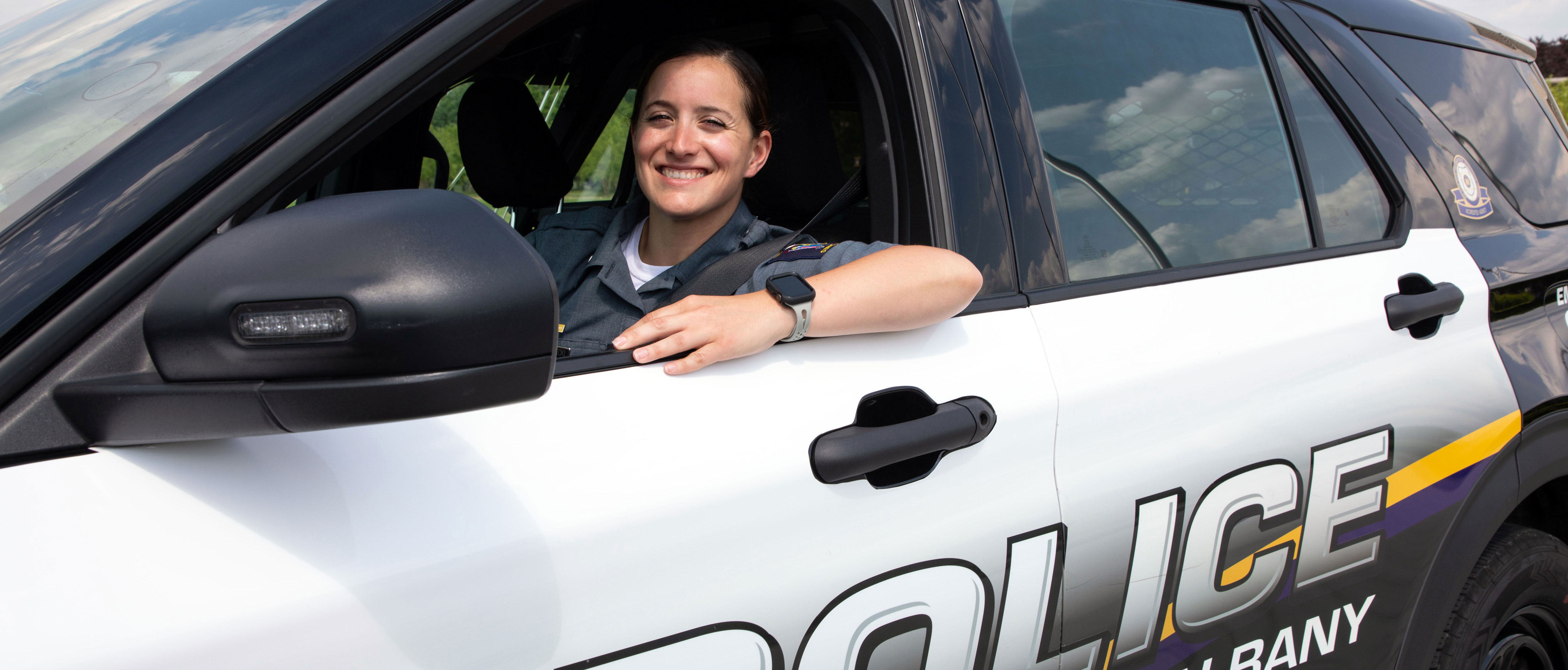 A smiling University police officer poses for a photo while seated inside her squad car.