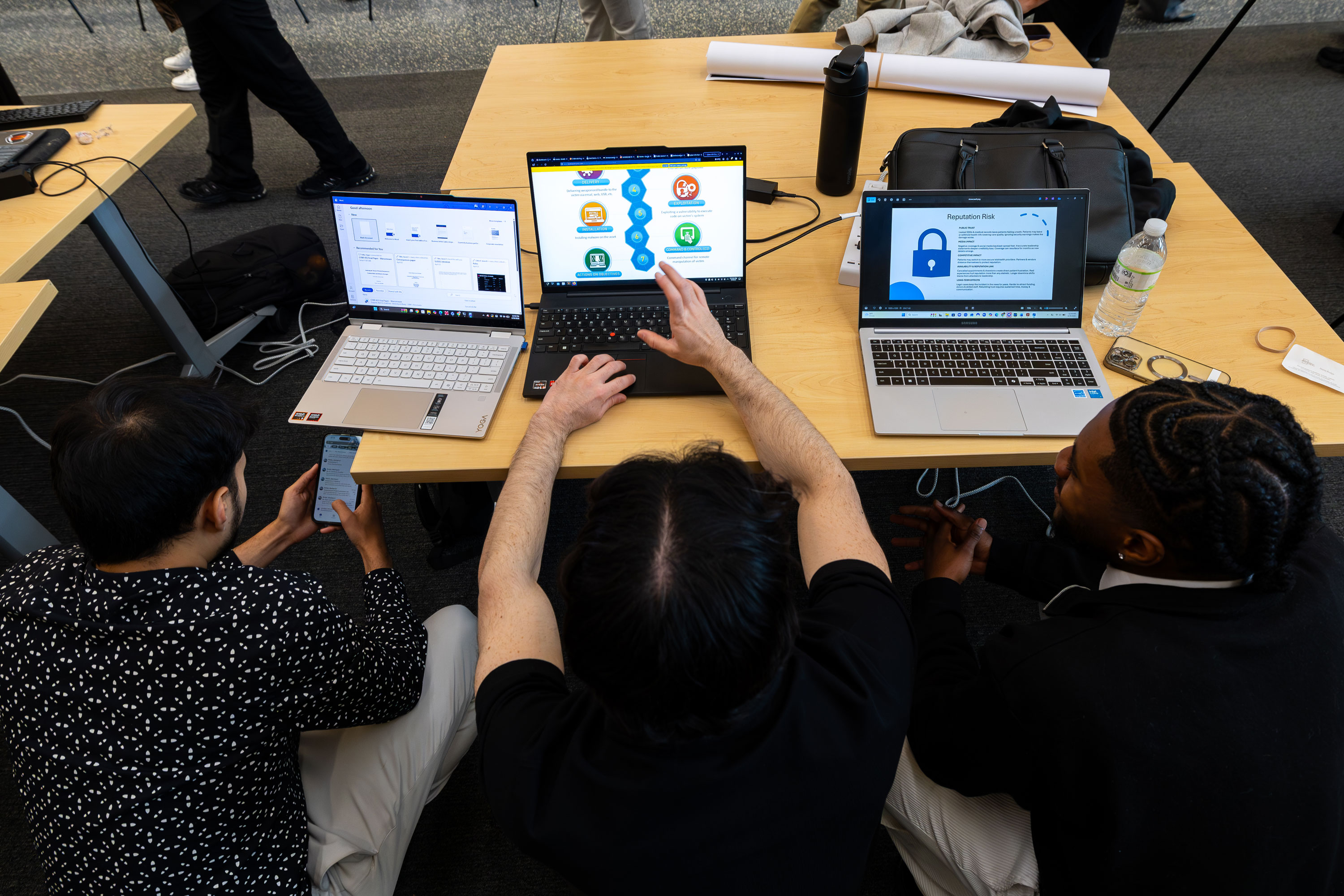 Three students sit at a table and collaborate on a presentation using their laptops.
