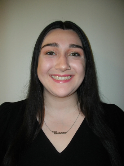 Portrait of Brianna Lennon, from the shoulders up. She is smiling and wearing a black V-neck top with a necklace bearing her first name. The background is gray. 