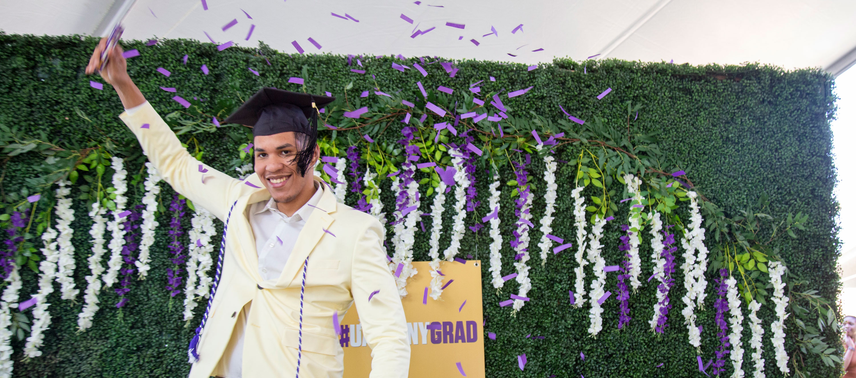 A student in a cream suit and black graduation cap raises one hand and smiles while posing for a photo under purple and white confetti.