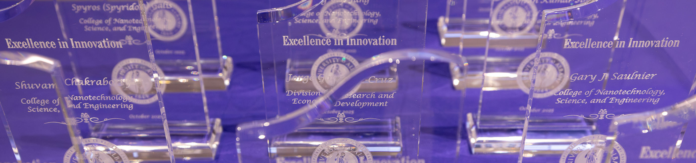 A dozen glass R&E Week awards are arranged in neat rows on a table with a purple tablecloth.