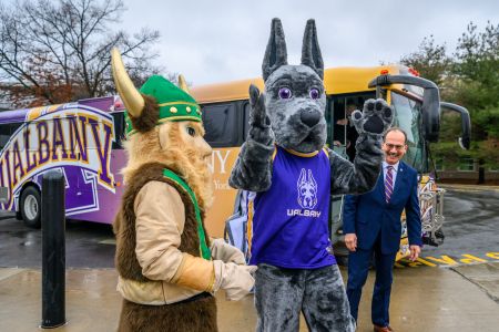 College mascots of a viking in a green helmet and a great dane in a blue shirt stand next to a smiling man in a suit in front of a bus painted with the word UAlbany.