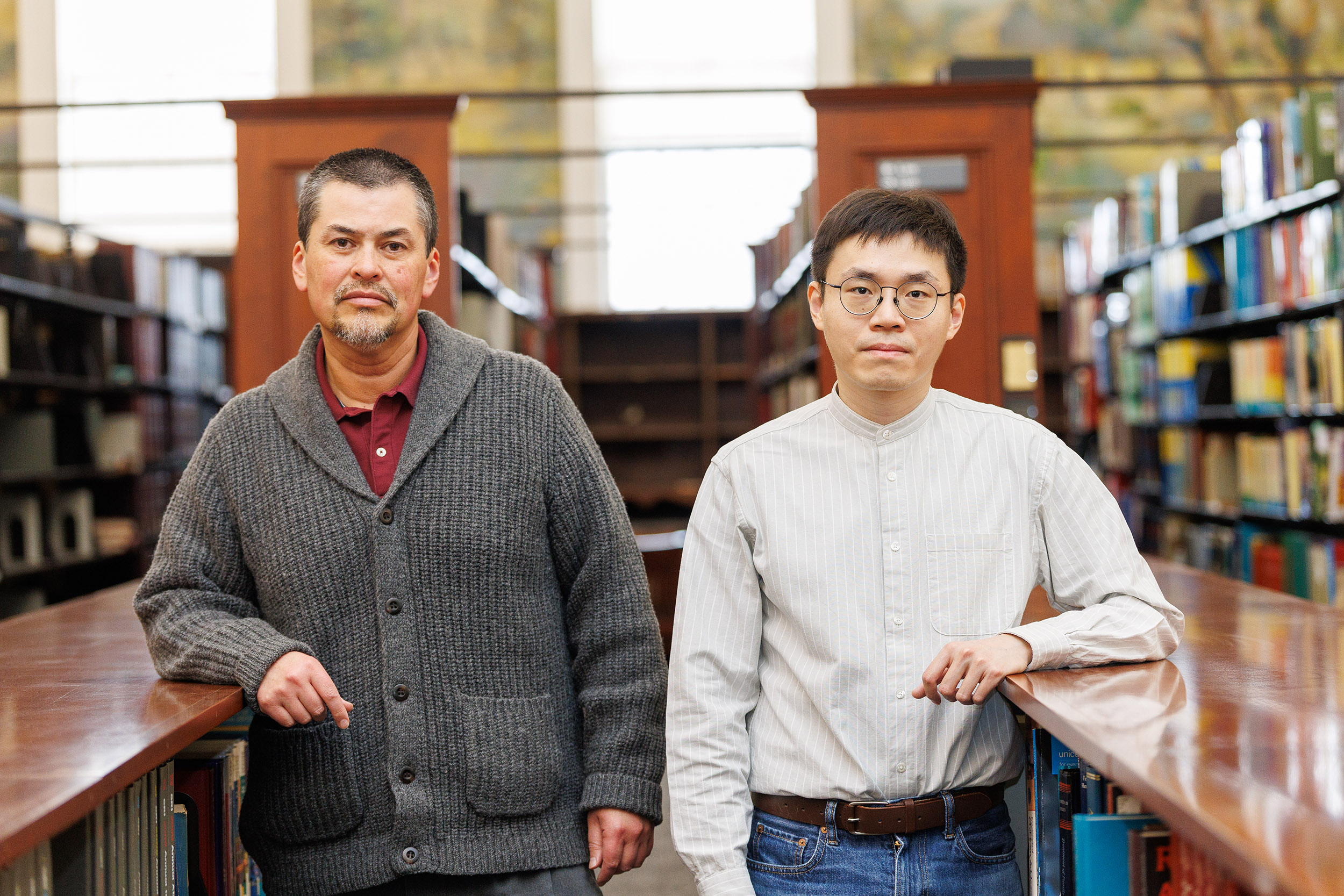 Two men stand amid bookshelves in a picturesque library facing forward with their elbows on the book shelves
