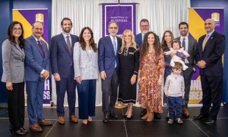 A line of smiling, dressed-up people, including a baby and a child pose before a banner reading "Massry School of Business"