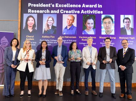 A line of faculty pose with glass trophies under a projection with their faces and the words" President's Excellence Award in Research and Creative Activities