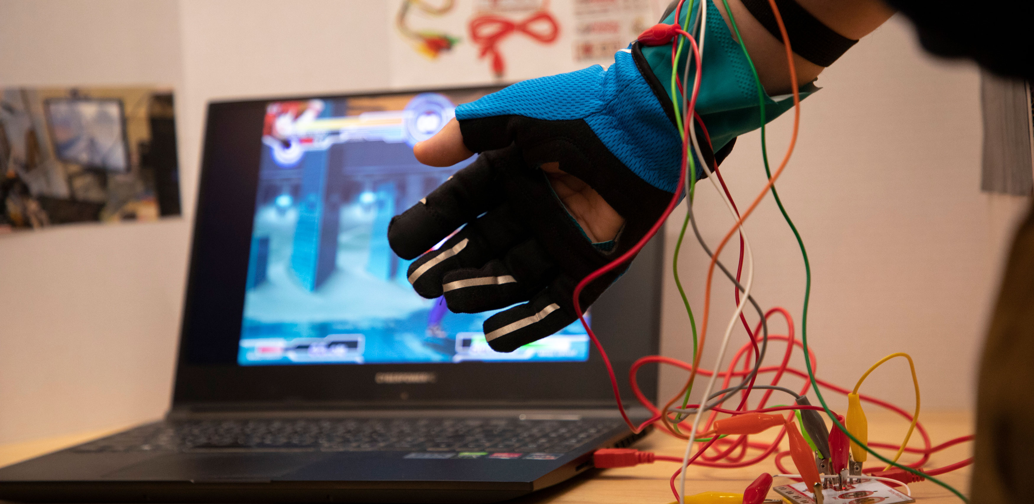 A hand demonstrates a wearable technology glove, connected to various colorful wires. A computer screen is visible in the background.