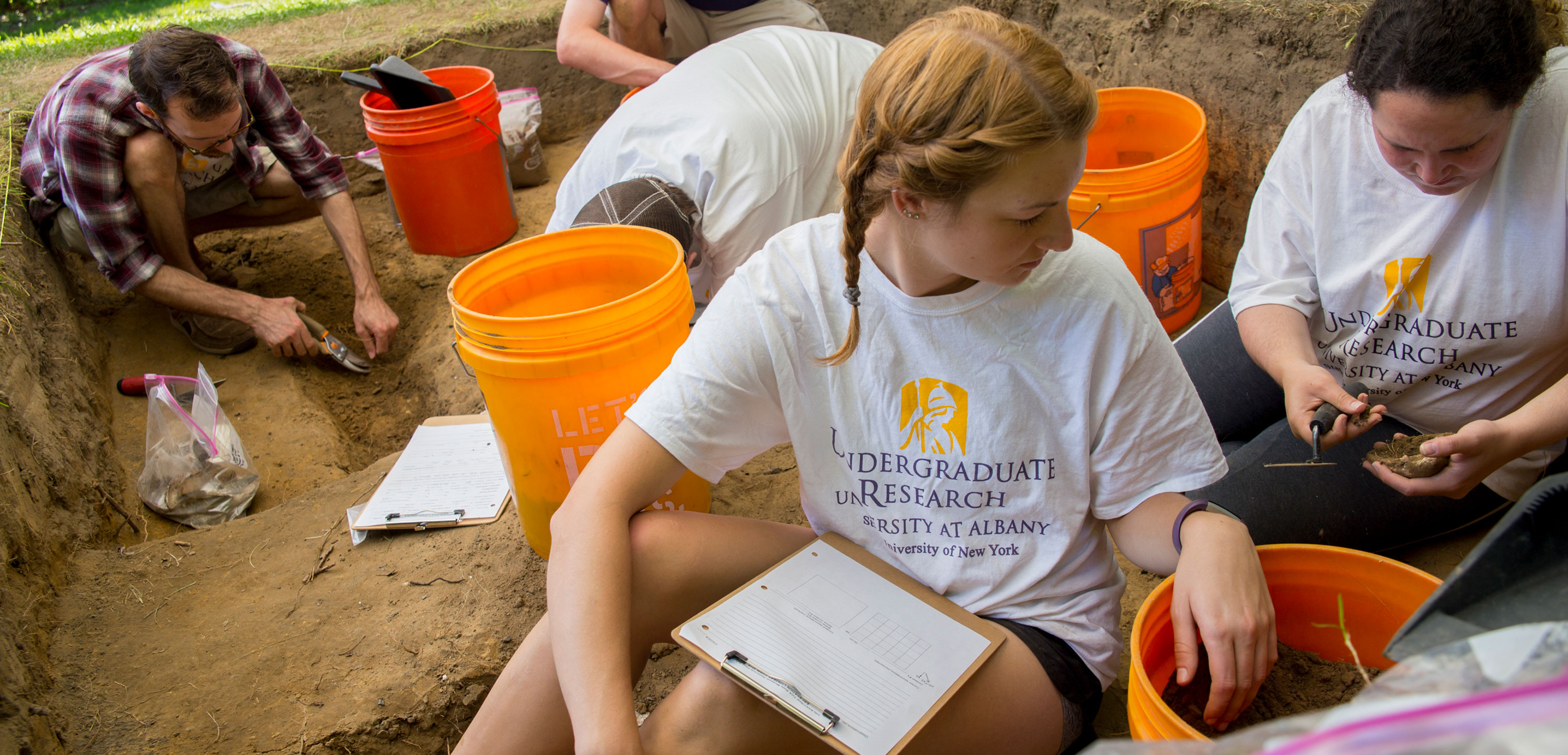 Students wearing Undergraduate Research, University at Albany, t-shirts sift through dirt at an outdoor archeological dig.