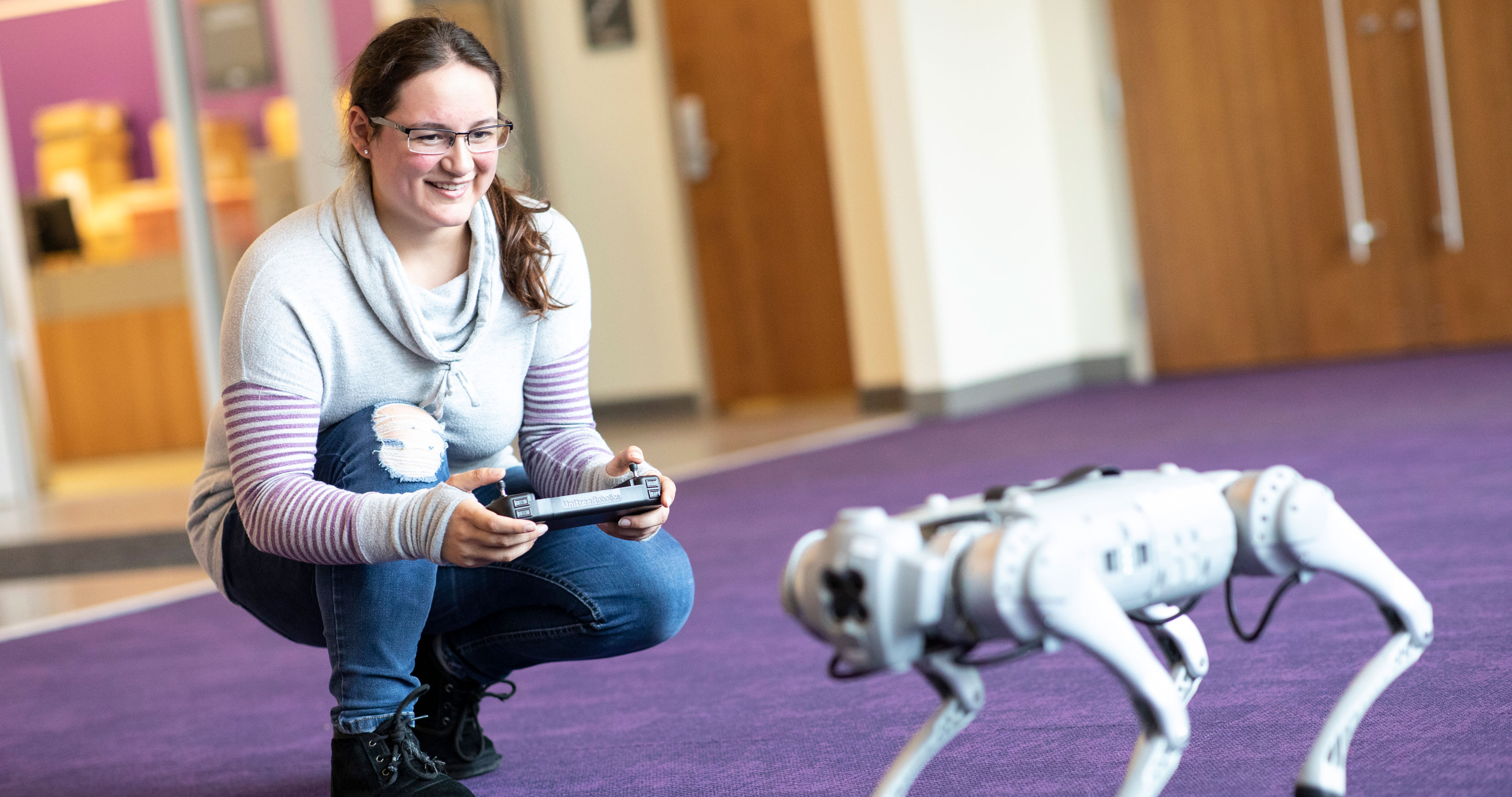 A smiling student holding a remote control crouches down next to a robot dog.