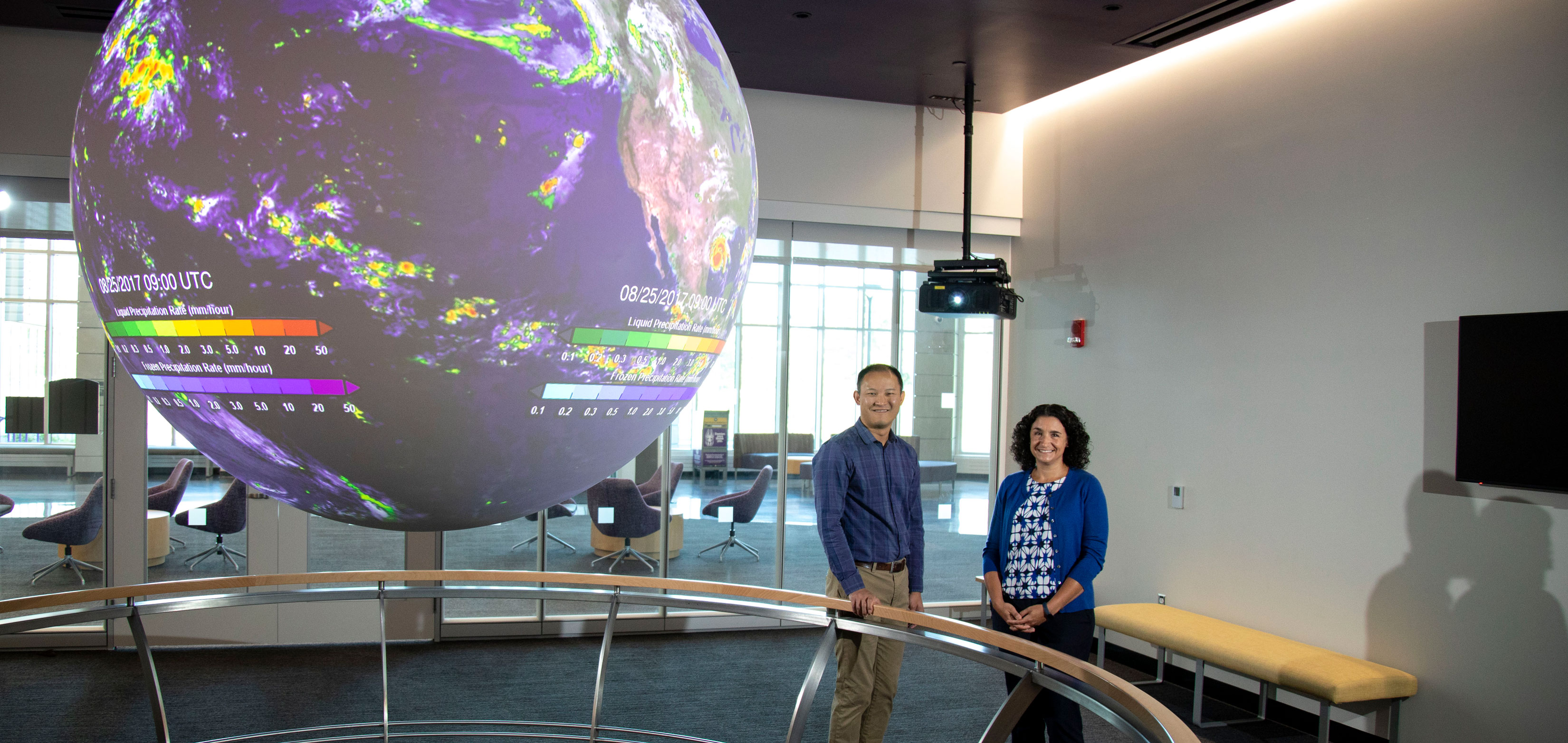 A man and a woman stand and smile for a camera in front of a huge digital globe showing live weather patterns.