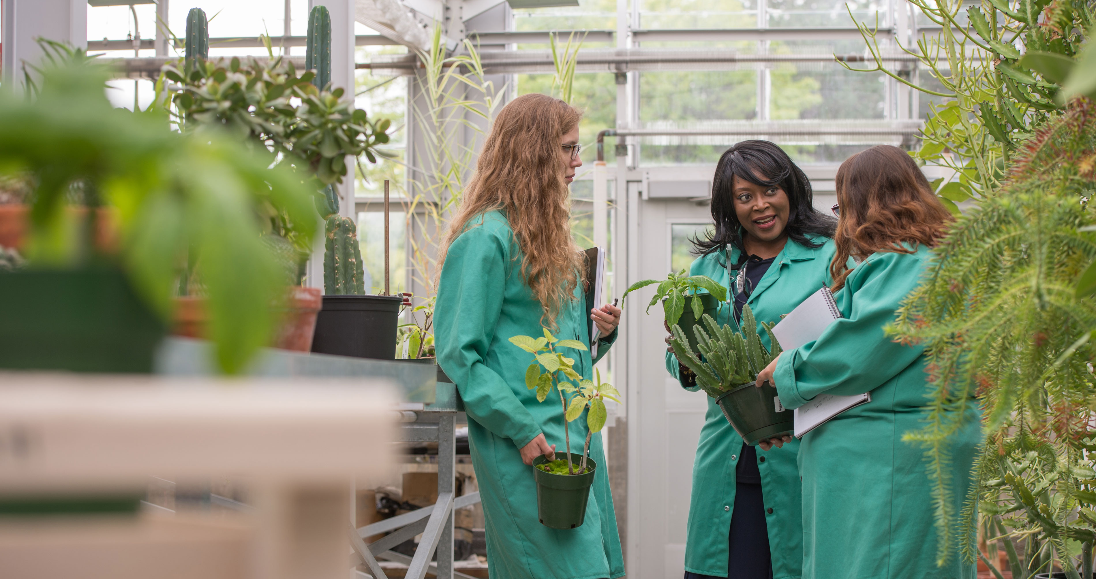 Three women in green lab coats holding plants and notebooks talk inside a greenhouse.