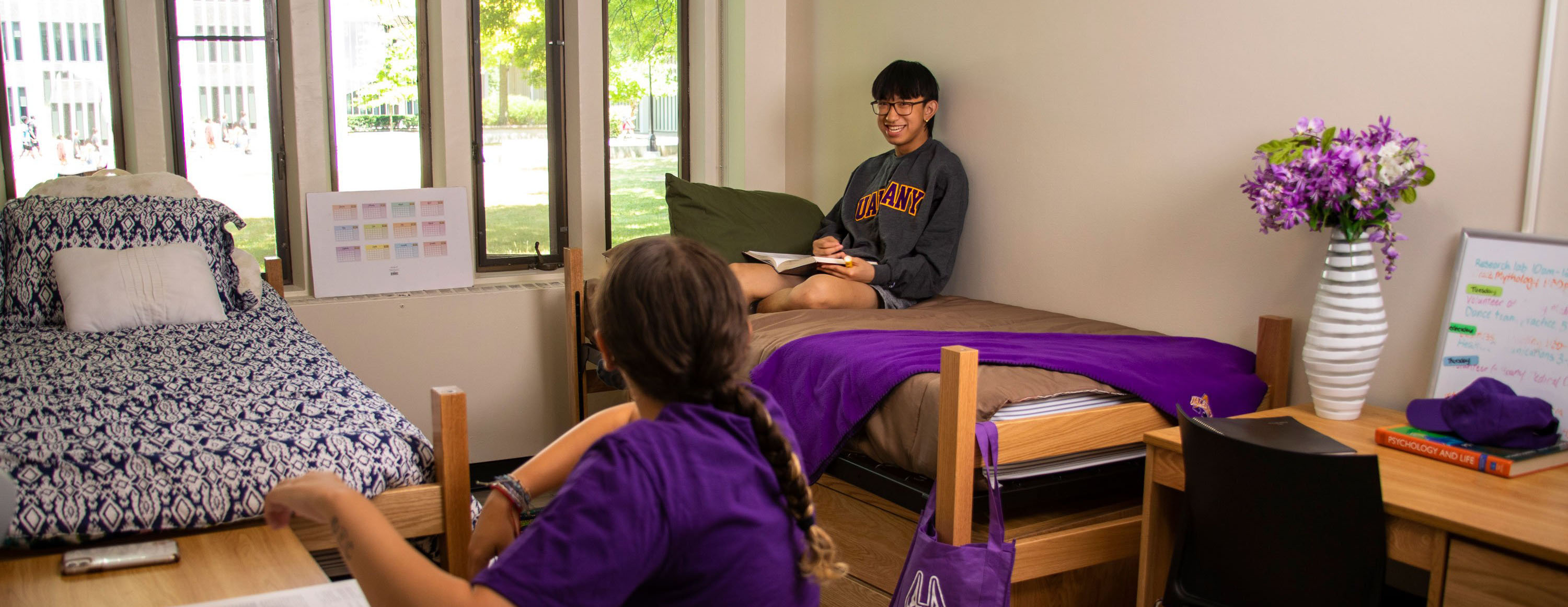 A double on-campus bedroom, with two twin beds, two desks and two chairs. One student sits at a desk and another sits on the bed, smiling and laughing together.