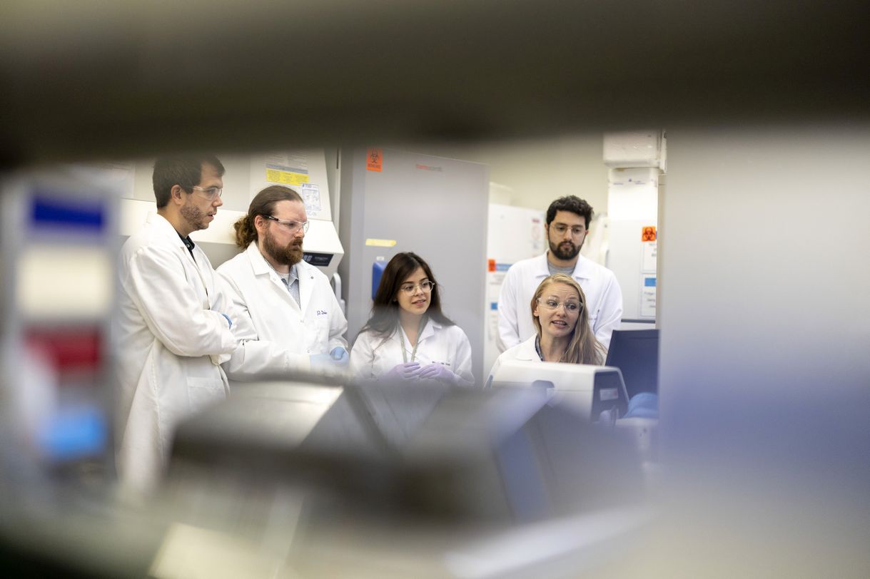 Peering through the space between lab shelving, five people wearing white lab coats focus their attention on a computer screen. 