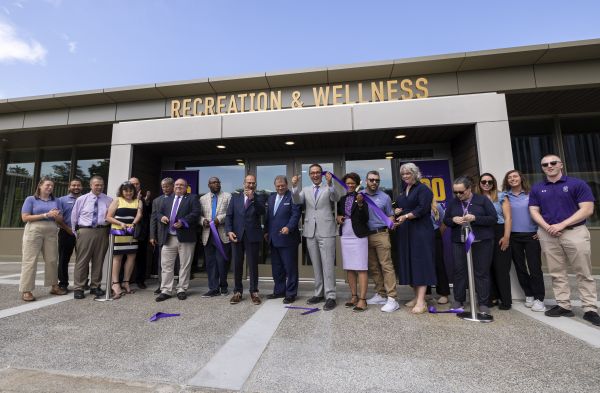 A line of smiling people cut a purple ribbon in front of a building with the words "Recreation & Wellness" above the doors.
