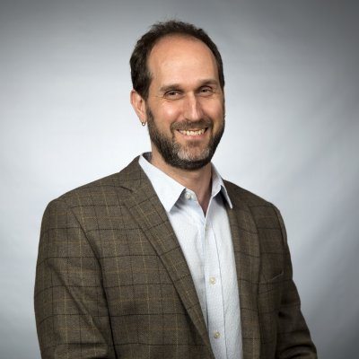A portrait of a a man with dark brown hair, a beard, wearing a brown tweed jacket and a collared shirt with the top button unbuttoned.