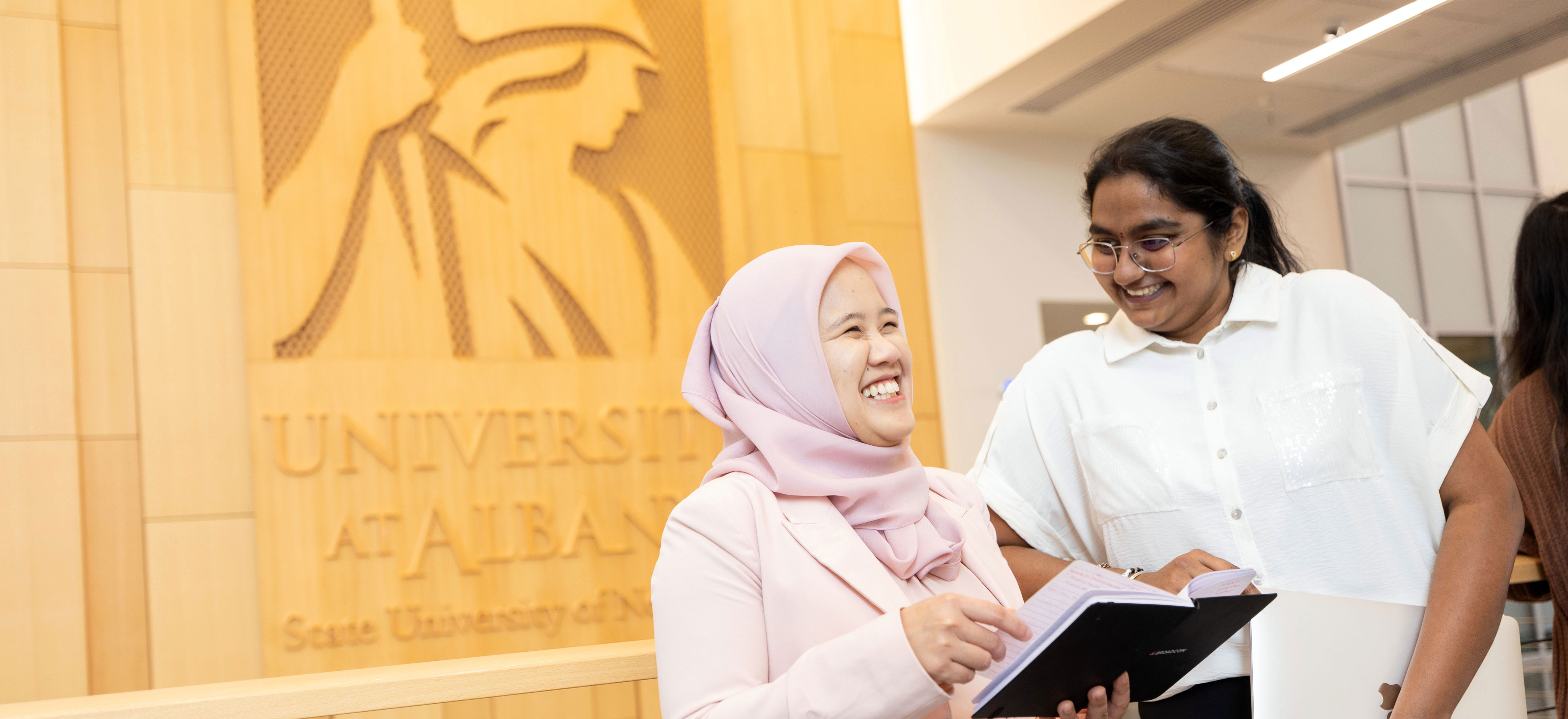 Two women smile as they talk inside the ETEC building. A wooden panel engraved with UAlbany's logo is behind them.