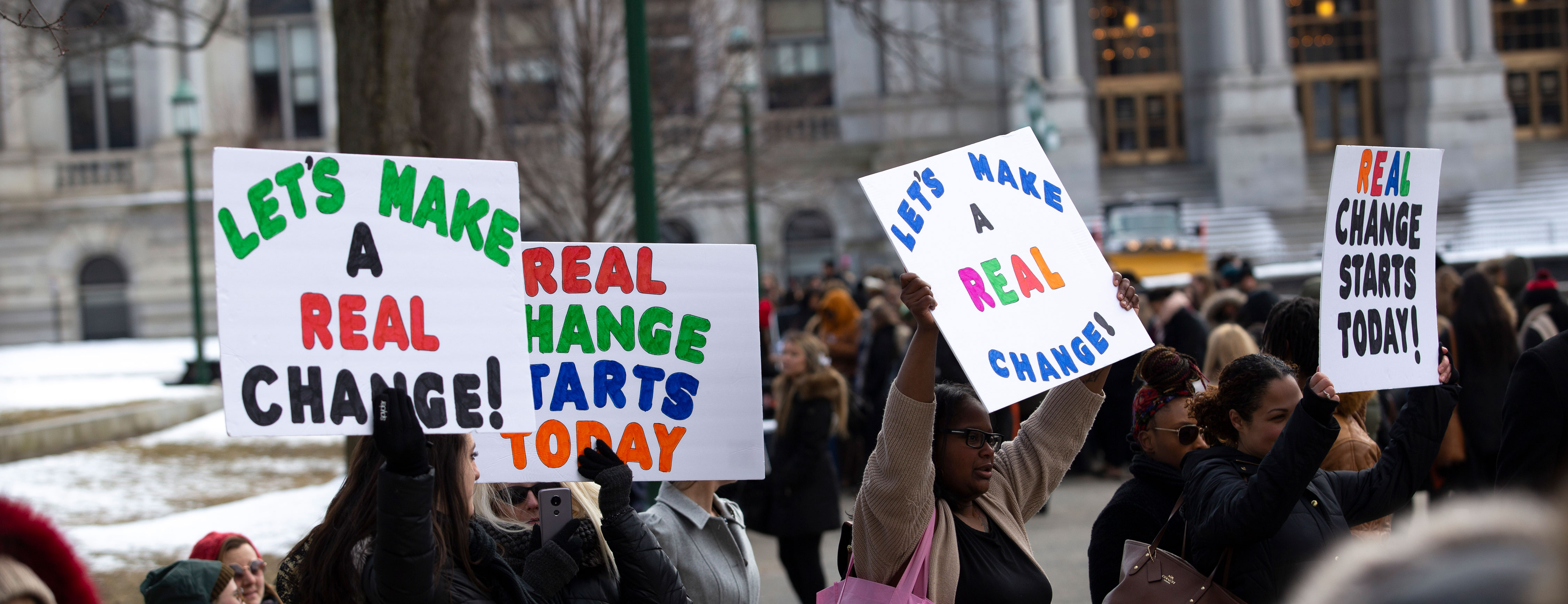 Social work students hold up posters that say, "Let's make real change," and "Real change starts today," at a gathering outside the New York State Capitol.