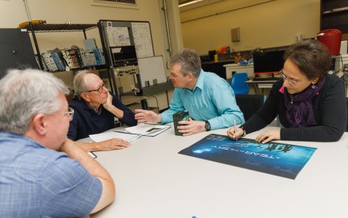 four people sit around a table, one of them signing a large blue poster