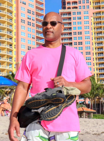 James is wearing a bright pink shirt and shades, standing on a beach with colorful high-rise buildings and a bright blue sky in the background.