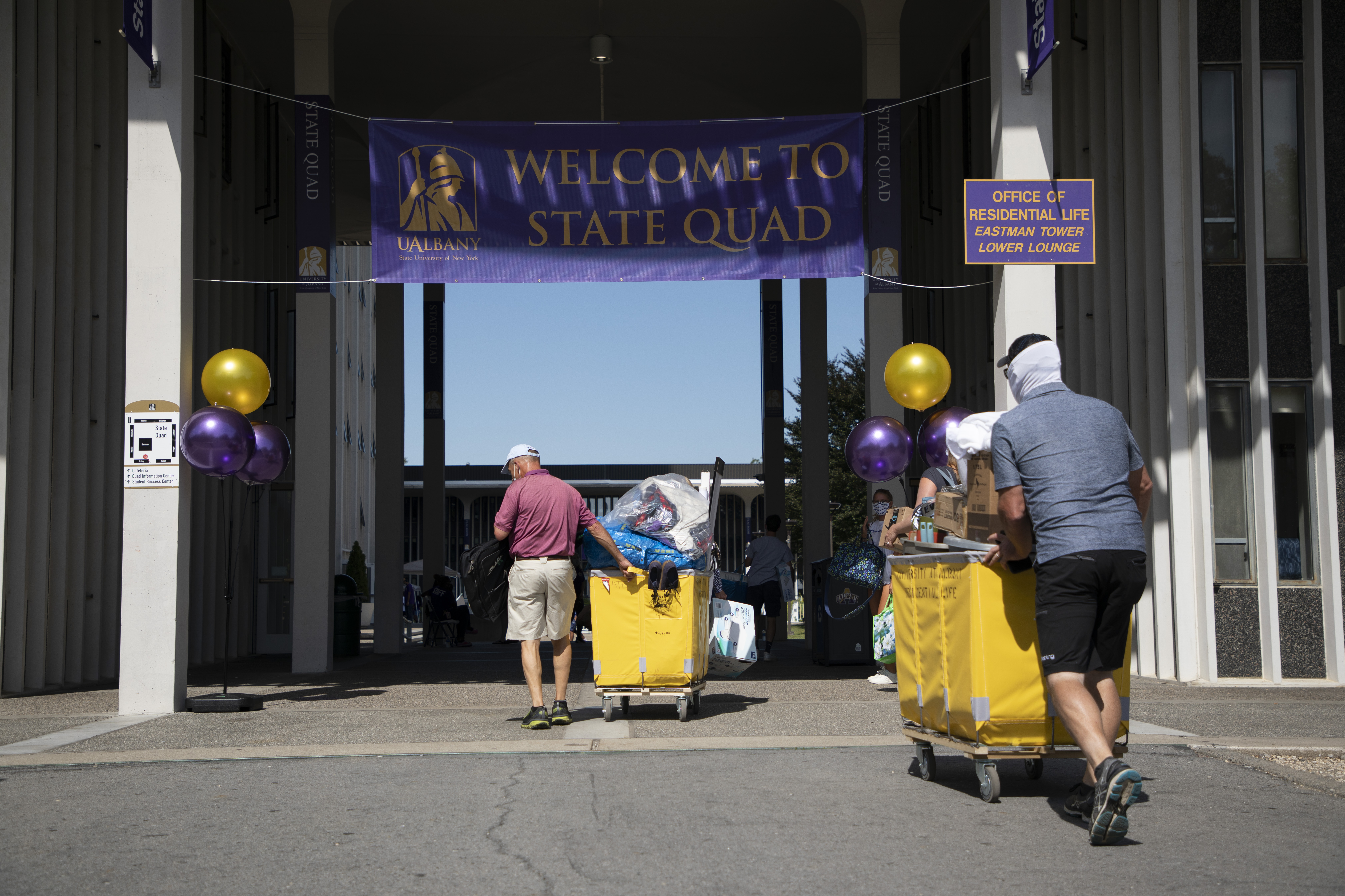 Two parents push yellow carts on move-in day, with a "Welcome to State Quad" banner visible
