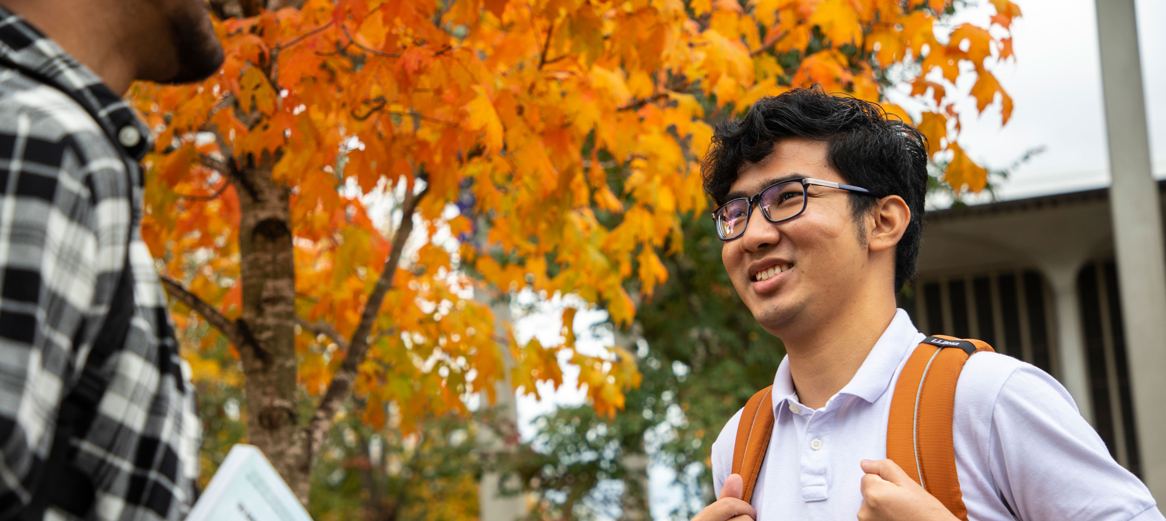 Two students smile as they talk in front of a tree with changing leaves.