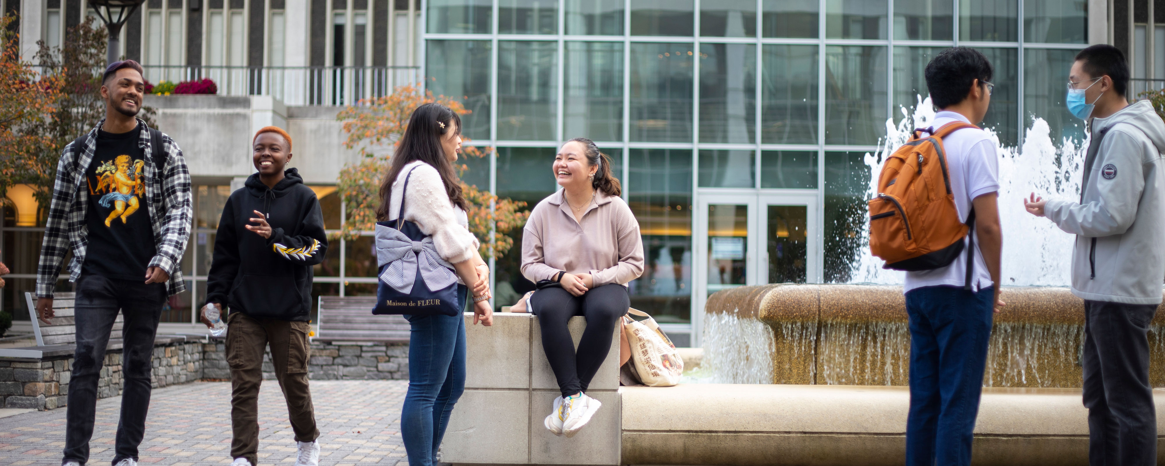 Six smiling students walk, sit and gather around a fountain on UAlbany's Uptown Campus.