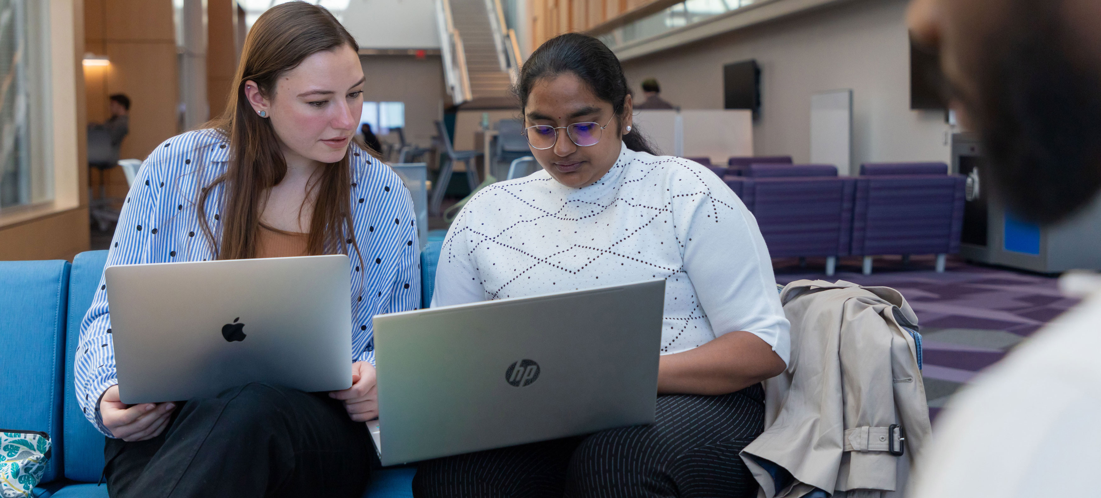 Two students sit on a couch and collaborate on their laptops.