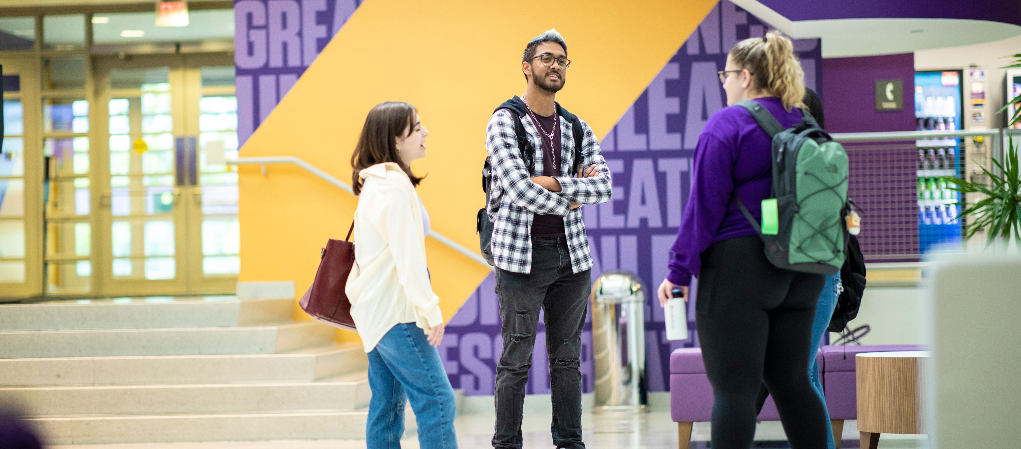 Three students with backpacks stand and speak in a UAlbany building's lobby.