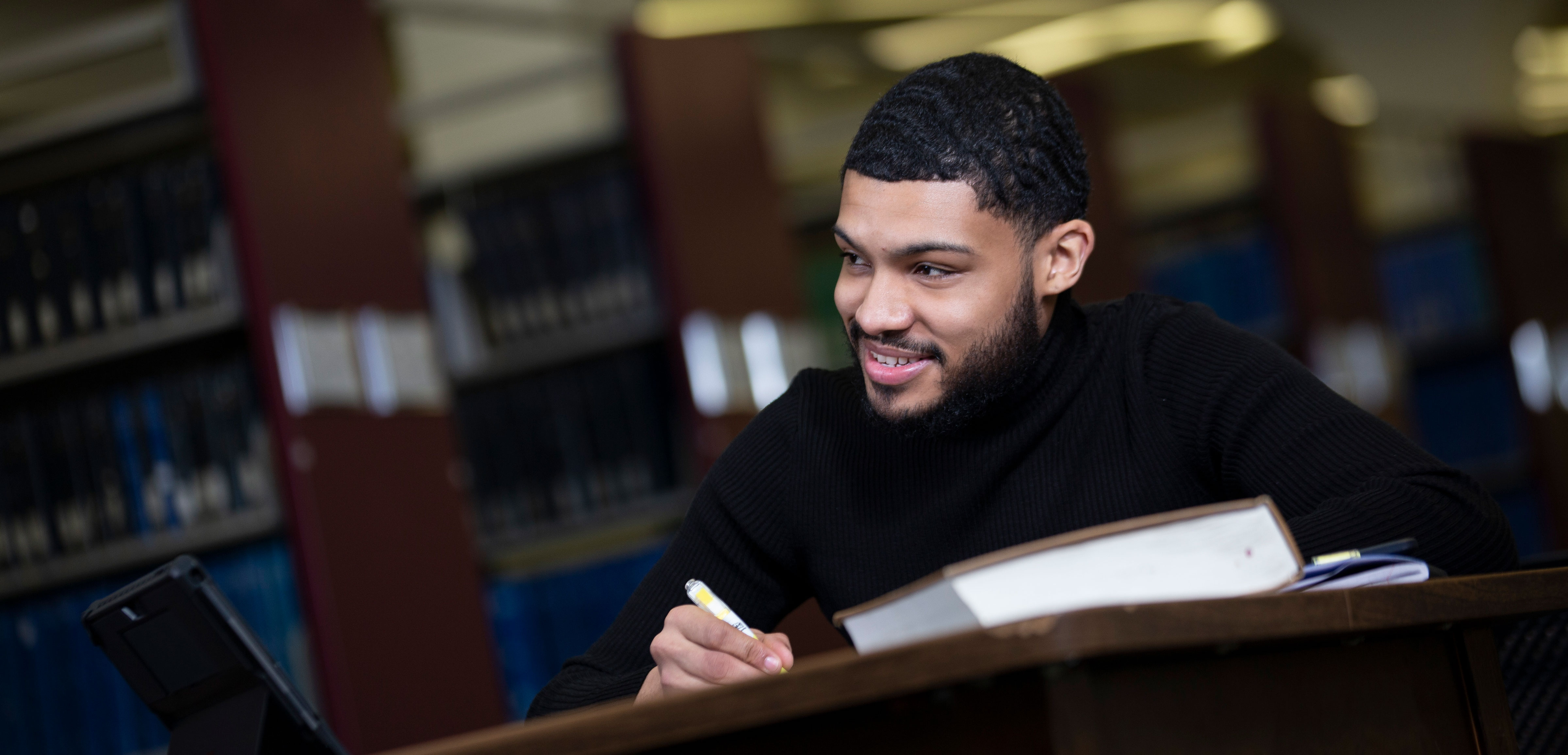 A smiling student sits at a desk in a library with notebooks and textbooks open on the table..